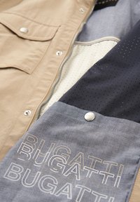 Beige Außenjackenstoff mit silbernem Metall, gefüttert mit marineblauem und hell gemustertem Stoff. Mit dem Text "BUGATTI" auf einer blauen Akzenttasche.