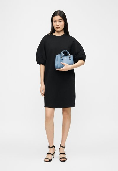 Γυναίκα με φαρδιά μαύρη φορεσιά και μαύρα τακούνια, κρατώντας ένα μικρό μπλε τσαντάκι, στέκεται μπροστά σε ένα απλό ανοιχτό γκρι φόντο.