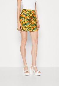 Mini falda floral con flores amarillas, naranjas y verdes sobre un fondo oscuro. Combinada con sandalias blancas de tacón cuadrado.