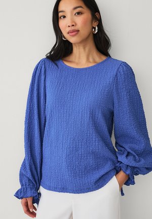 Ung kvinde med langt mørkt hår iført sølvfarvede øreringe og en tekstureret blå bluse med lange ærmer og opsmøgede manchetter samt hvide bukser.