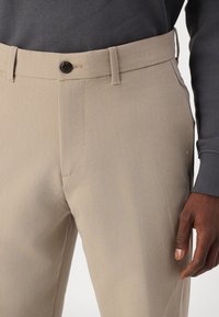 Bež hlače iz teksturirane tkanine, z gumbu zaprtjem, stranskimi žepi in slim fit krojem. V kombinaciji z temno sivo majico s kapuco.