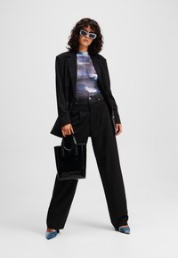 Zwarte blazer over een blauw en grijs gestippeld topje, brede zwarte broek, en blauwe puntige schoenen, terwijl ze een kleine zwart glanzende handtas vasthoudt.