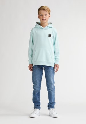 Sweat à capuche bleu clair avec poche avant et écusson logo noir, associé à un jean bleu et des baskets blanches, posé devant un fond uni.