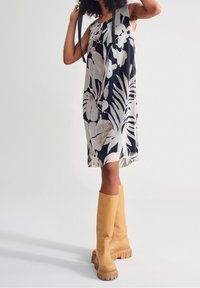 Blumenkleid mit schwarzem Grund und großen weißen und beigen Pflanzenmustern, ergänzt durch knöchelhöhe gelbe Plateau-Stiefel.