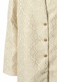Beige stof met een patroon van geometrische vormen. Drukknoopsluitingen met tekstuurlijke goudaccenten. Glad, licht glanzende textuur.