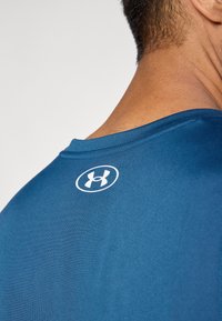 Maglietta sportiva a maniche corte di colore blu scuro con una texture liscia. Presenta un logo bianco nella parte superiore della schiena, collo rotondo con dettagli di cuciture pulite.