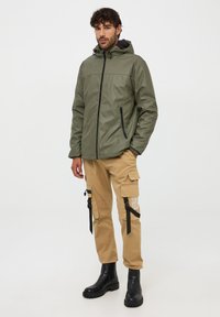 Giacca impermeabile verde oliva con cerniera nera, cappuccio regolabile e tasche laterali. Abbinata a pantaloni cargo beige e stivali neri robusti.