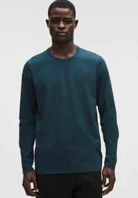 Dlhý rukáv zelená bavlnená košeľa s okrúhlym výstrihom, hladkou textúrou a priliehavým strihom. Nosí sa s čiernymi nohavicami, minimalistický štýl.