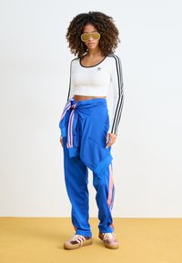 Long-sleeve white crop top with black stripes, blue sweatpants with pink accents, and oversized yellow sunglasses. Textured fabric finishes.