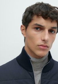 Tmavě námořnická bunda na zip s vysokým límcem, vrstvená přes světle šedý rolák. Hladká textura látky, úzký střih a bez viditelných vzorů.