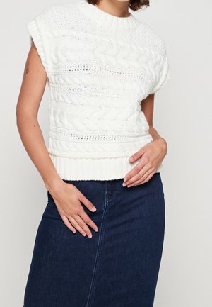 Kobieta w białym bezrękawniku z dzianiny warkoczowej, połączonym z ciemnoniebieską spódnicą dżinsową, z rękami opartymi na biodrach.