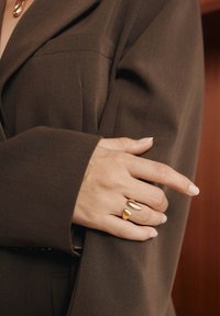 Zlatý prsteň s vrstveným, slzovitým dizajnom, nosený na ruke, dopĺňajúci tmavohnedý oversize kabát. Hladký povrch, minimálne detaily.