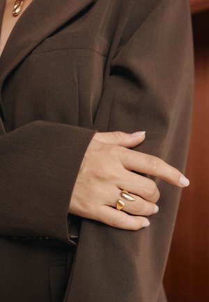 Kuldne sõrmus kihilise, tilgakujulise disainiga, kantud käel, täiustades tumepruuni üle mõõdu suuruse jakki. Sile tekstuur, minimaalne detailide arv.