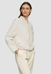 Veste bomber beige clair avec un col rond, une fermeture éclair à l'avant et une poche latérale. Portée sur un pantalon assorti beige clair. Tissu lisse et léger.