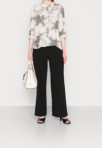 Blusa chiara con motivo a pois neri, con bottoni sul davanti e maniche arrotolate; abbinata a pantaloni larghi neri e una borsa beige. Sandali neri con tacco.