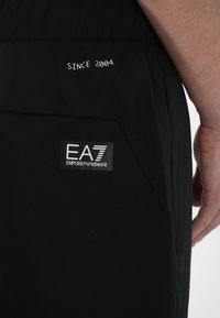 Juodi sportiniai šortai su glotnia tekstūra, su išsiuvinėtu užrašu "Nuo 2004" ir EA7 Emporio Armani logotipo pleistru šone.