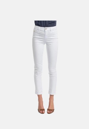 Donna che indossa jeans bianchi slim-fit e tacchi aperti marroni, abbinati a un top scuro infilato, su uno sfondo chiaro e uniforme.
