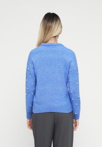 Kobieta z długimi blond włosami, ubrana w niebieski sweter z perłowymi zdobieniami na rękawach i ciemnoszare spodnie, widoczna od tyłu.