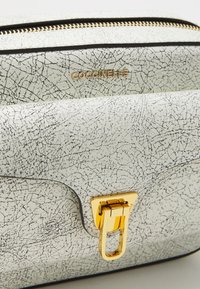 Borsa in pelle bianca testurizzata con finitura screpolata, hardware dorato e chiusura a lucchetto. Dettaglio della cerniera lungo i bordi superiori.