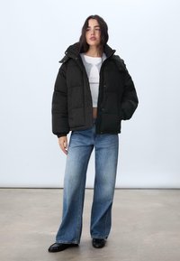 Giubbino imbottito nero con chiusura a zip e cappuccio, dotato di tasche laterali e fitting ampio. Indossato sopra una maglietta bianca e abbinato a jeans a gamba larga.