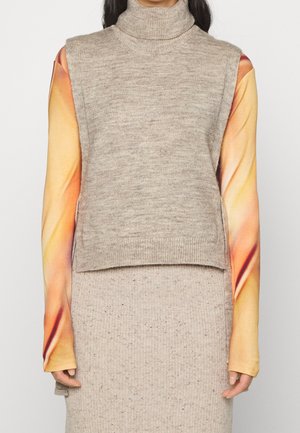 Personen bærer en beige ærmeløs rullekrave sweater over en langærmet top med orange og gul gradientærmer.