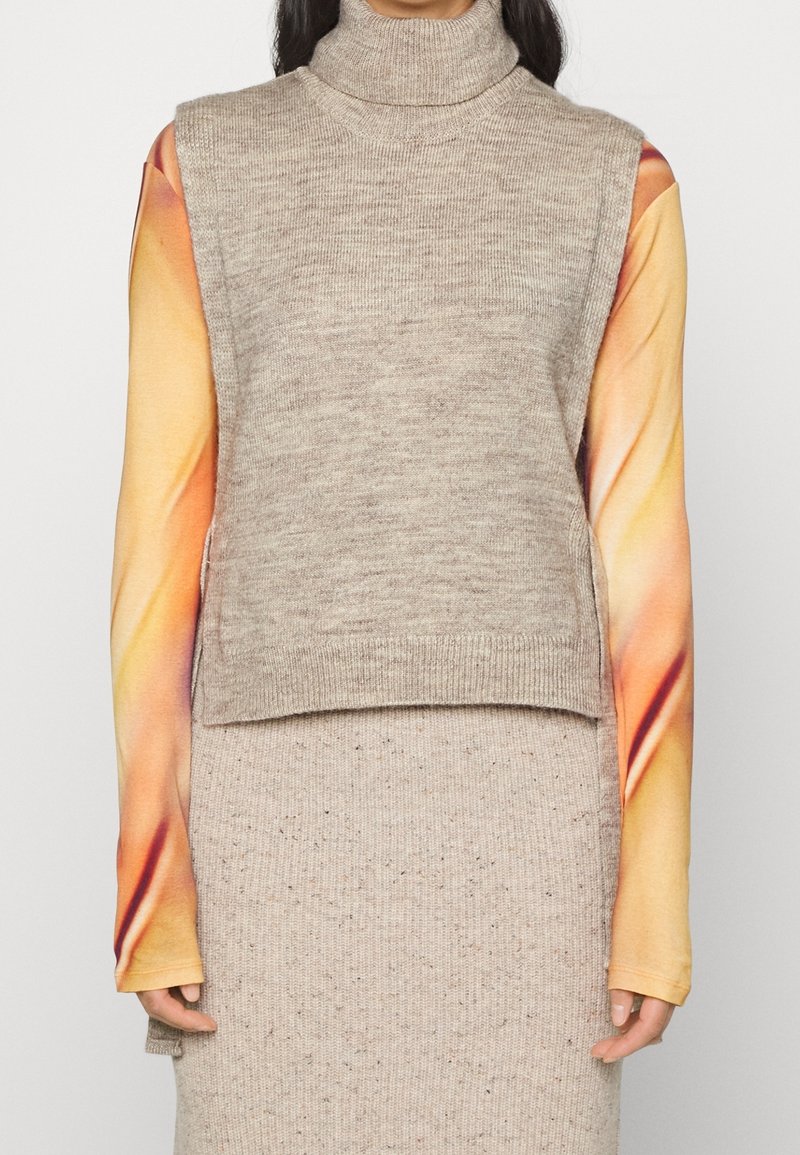 La personne porte un pull beige sans manches à col roulé par-dessus un haut à manches longues avec des manches dégradées orange et jaune.