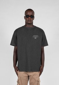Mørkegrå bomulds t-shirt med korte ærmer, rund halsudskæring og lille hvid tekst på brystet. Bærende med beige cargo shorts og solbriller.