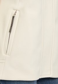 Hvid fleecejakke med en lynlåslomme, flade sømme og en struktureret overflade. Stoffet er blødt og glat mod huden.