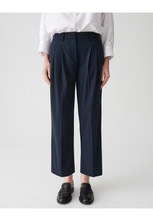 Pantalons plissés navy en tissu lisse, avec une coupe droite et des poches latérales, associés à des mocassins noirs et une chemise blanche.