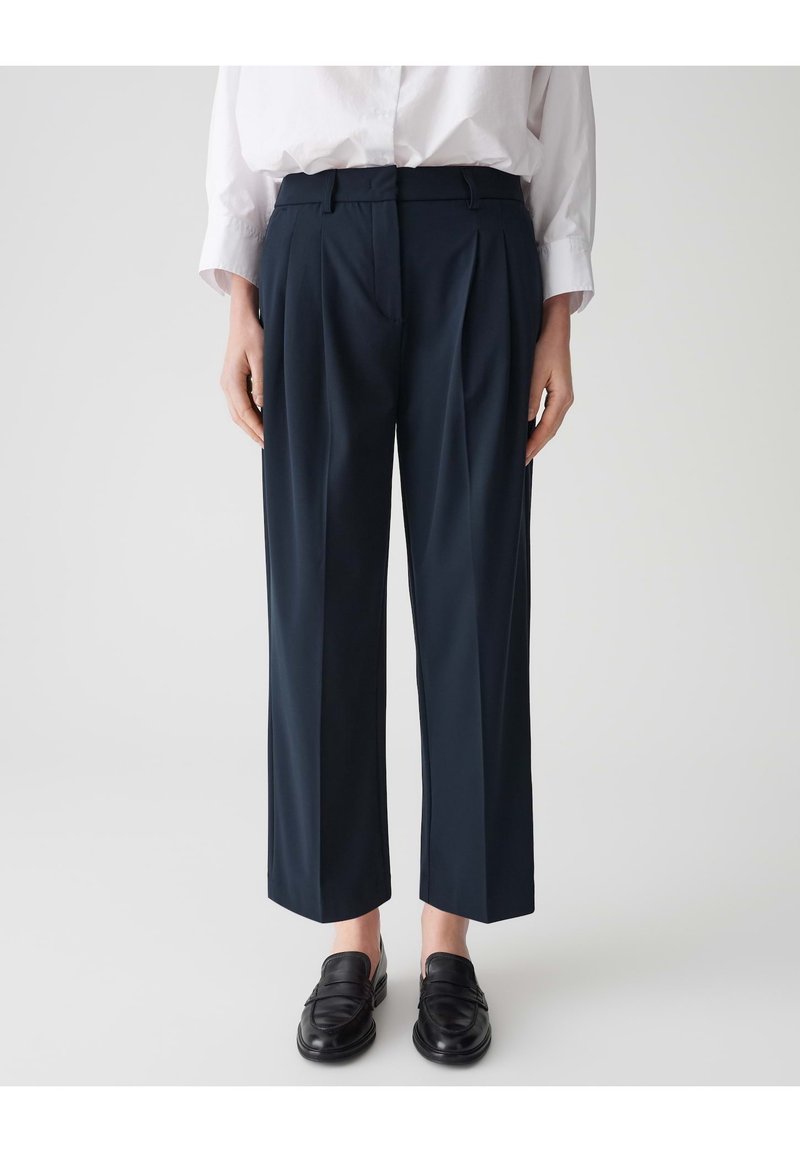 Marineblauwe geplooide broek van een gladde stof, met een rechte snit en zijzakken, gestyled met zwarte loafers en een wit shirt.