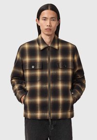 Giacca a quadri nera e marrone con zip frontale, due tasche sul petto con bottone e colletto, posizionata su uno sfondo neutro.