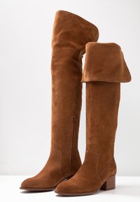 Bottes hauteur genou en daim marron avec un revers plié, fermeture éclair latérale et talon bloc, posées sur une surface et un fond blancs.