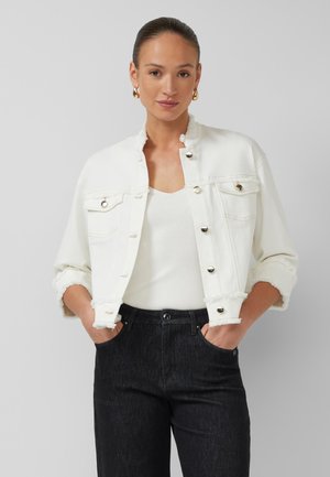 Frau mit zurückgekämmtem Haar trägt goldene Creolen, weiße kurze Knopfjacke, weißes Tanktop und schwarze High-Waist-Jeans mit Händen in den Taschen.