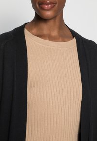 Cárdigan negro sobre un top beige de canalé. El cárdigan presenta una textura suave con diseño de frente abierto y escote ligeramente redondeado en la parte superior.