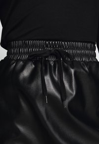 Svart fusk läderkjol med en rynkad elastisk midja och dragsko. Slät textur med metalliska accenter på dragskoändarna.
