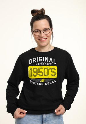 Donna sorridente con occhiali che indossa una felpa nera con la scritta "Originale e non restaurato, prodotti vintage degli anni '50" e jeans azzurri.
