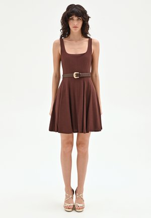 Femme aux cheveux bouclés et foncés, portant une robe marron sans manches avec une ceinture et des sandales à talons hauts dorées à brides, se tenant devant un fond blanc.