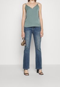 Vihreä hihaton paita, jossa on aaltoileva kaulus, yhdistettynä leveälahkeisiin sinisiin farkkuihin ja mustiin sandaaleihin, joissa on remmit. Pienestä käsilaukusta pidetään toisessa kädessä.