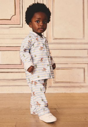 Małe dziecko z kręconymi włosami w niebiesko-białych pasiastych piżamkach z nadrukiem kowbojów i kaktusów, stojące na drewnianej podłodze na tle jasnej, lamperyjowej ściany.