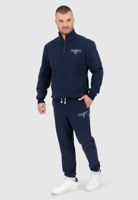 Pullover in pile blu navy con colletto alto e zip, abbinato a pantaloni joggers in pile coordinati. Presenta dettagli ricamati e lacci bianchi.