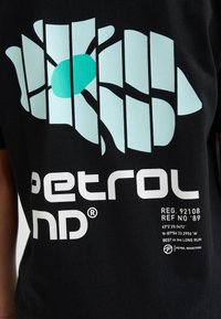 Schwarzes T-Shirt mit einem hellblauen Grafikdesign, das einen zentralen Kreis, sich kreuzende Linien und weißen Text mit Marken- und Registrierungsinformationen zeigt.