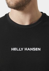 Camiseta de algodón negra con cuello redondo y texto bordado en blanco "HELLY HANSEN" en la parte frontal. Textura suave, diseño casual.