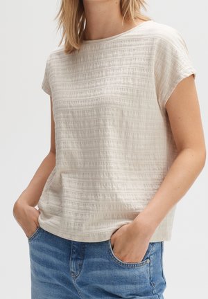 Woman wearing short-sleeve textured beige top with round neckline and blue denim jeans with hands in pockets against plain background.