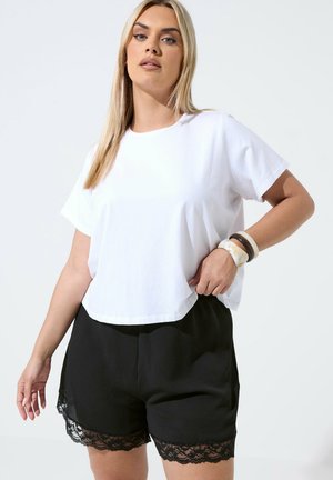 Kvinde med langt blondt hår iført en hvid kortærmet skjorte, sorte shorts med blonderkant og tre armbånd på højre håndled.