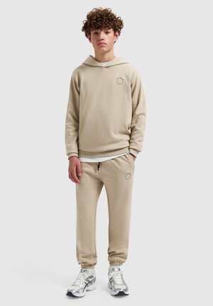 Teenager mit lockigem Haar, der einen beigen Hoodie und passende Jogginghosen trägt, steht mit einer Hand in der Tasche vor einem einfarbigen Hintergrund.