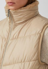 Beige gewatteerde vest met hoge kraag en zichtbare rits, gedragen over een gebreide beige trui door een persoon.