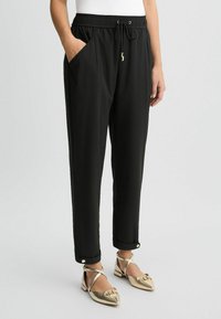 Schwarze, taillierte Hosen mit elastischem Bund, Seitentaschen und goldenen Hardware-Details an den Bündchen. Glattes, leichtes Material.