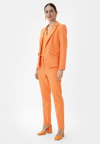 Oranger maßgeschneiderter Anzug mit Blazer und eng anliegenden Hosen. Glänzende orangefarbene Blockabsatzschuhe. Glattes Material und strukturierte Form.