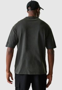Homme portant un t-shirt oversize vert foncé, un pantalon cargo noir et une casquette de baseball noire, tourné le dos contre un fond uni.