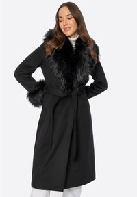 Schwarzer Mantel mit einem Kragen und Manschetten aus Kunstpelz, tailliertem Design, langen Ärmeln und glatter Textur. Die Gesamtform ist figurbetont und elegant.